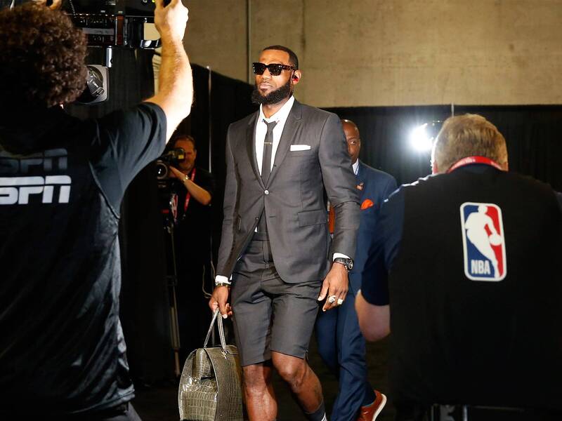 Lebron leaving a basketball arena wearing sunglasses and a Thom Browne shorts suit and carrying a grey leather bag.