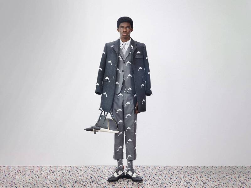 Man wearing a grey suit and overcoat and saddle shoes, holding a dolphin handbag. The suit, coat, and socks have a white dolphin pattern on them. From the Spring 2020 Mens Look