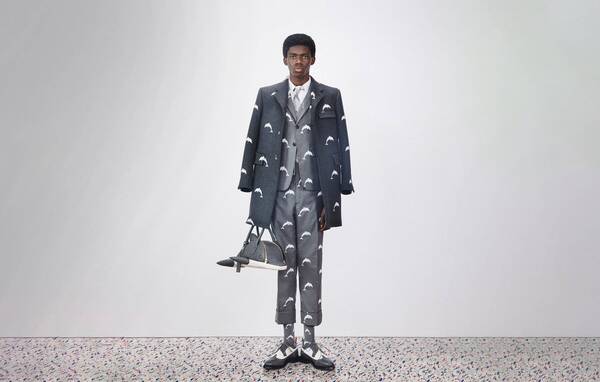 Man wearing a grey suit and overcoat and saddle shoes, holding a dolphin handbag. The suit, coat, and socks have a white dolphin pattern on them. From the Spring 2020 Mens Look