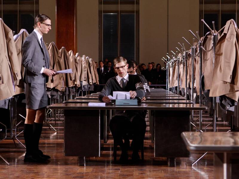 Thom Browne fashion show with models in shorts suits seated at rows of desks with tan coats hanging on coat hangers next to the desks. 2009 Pitti Uomo