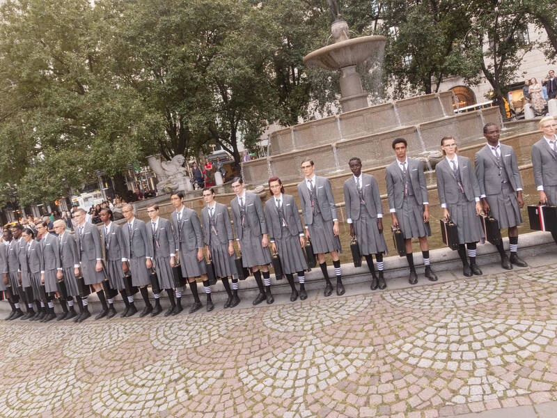 Models wearing grey Thom Browne suits and skirts, carrying briefcases stand in front of a fountain for Vogue