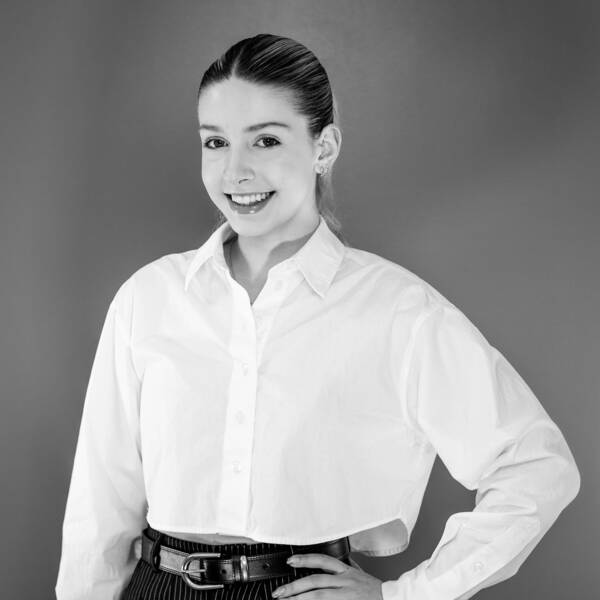 Taylor Dellelce on a black and white background, with a white blouse and belt, smiling