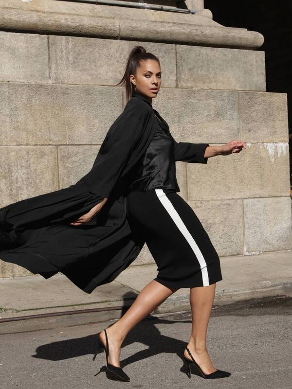 Julie Henderson model picture walking in black high heals, stone background, black long waving jacket, a black skirt with a white stripe, hair in pony tail